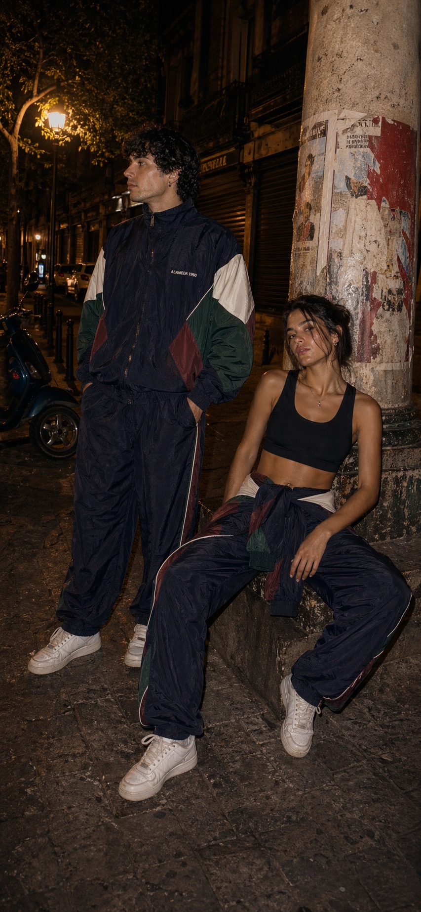 Man in a full Alameda 1990 tracksuit and woman with the tracksuit jacket tied at her waist over a black gym top beside an old column at night.