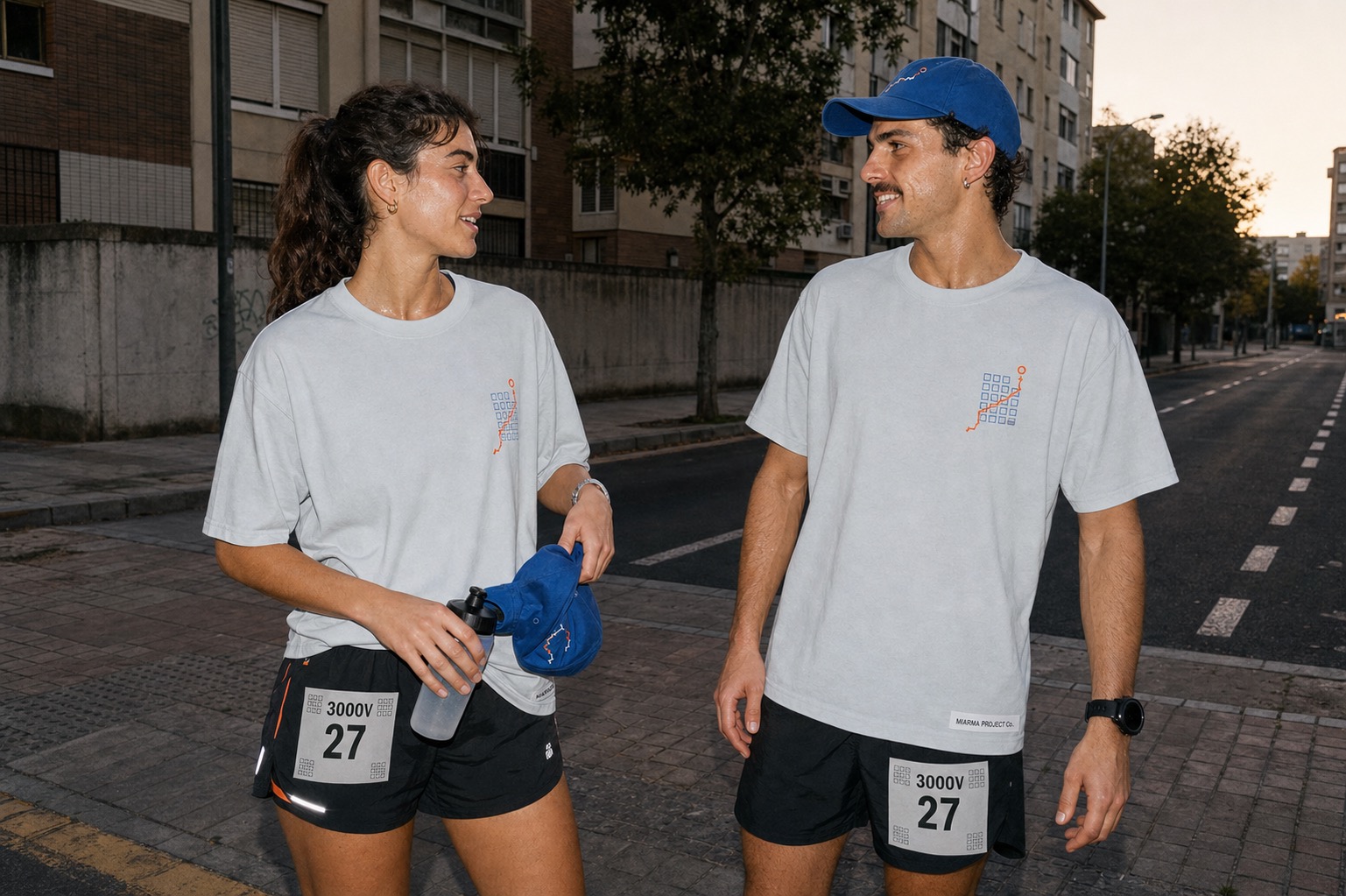 Two adult runners wearing ice-gray running club tees with cobalt and safety-orange details near Sevilla residential blocks at dawn.