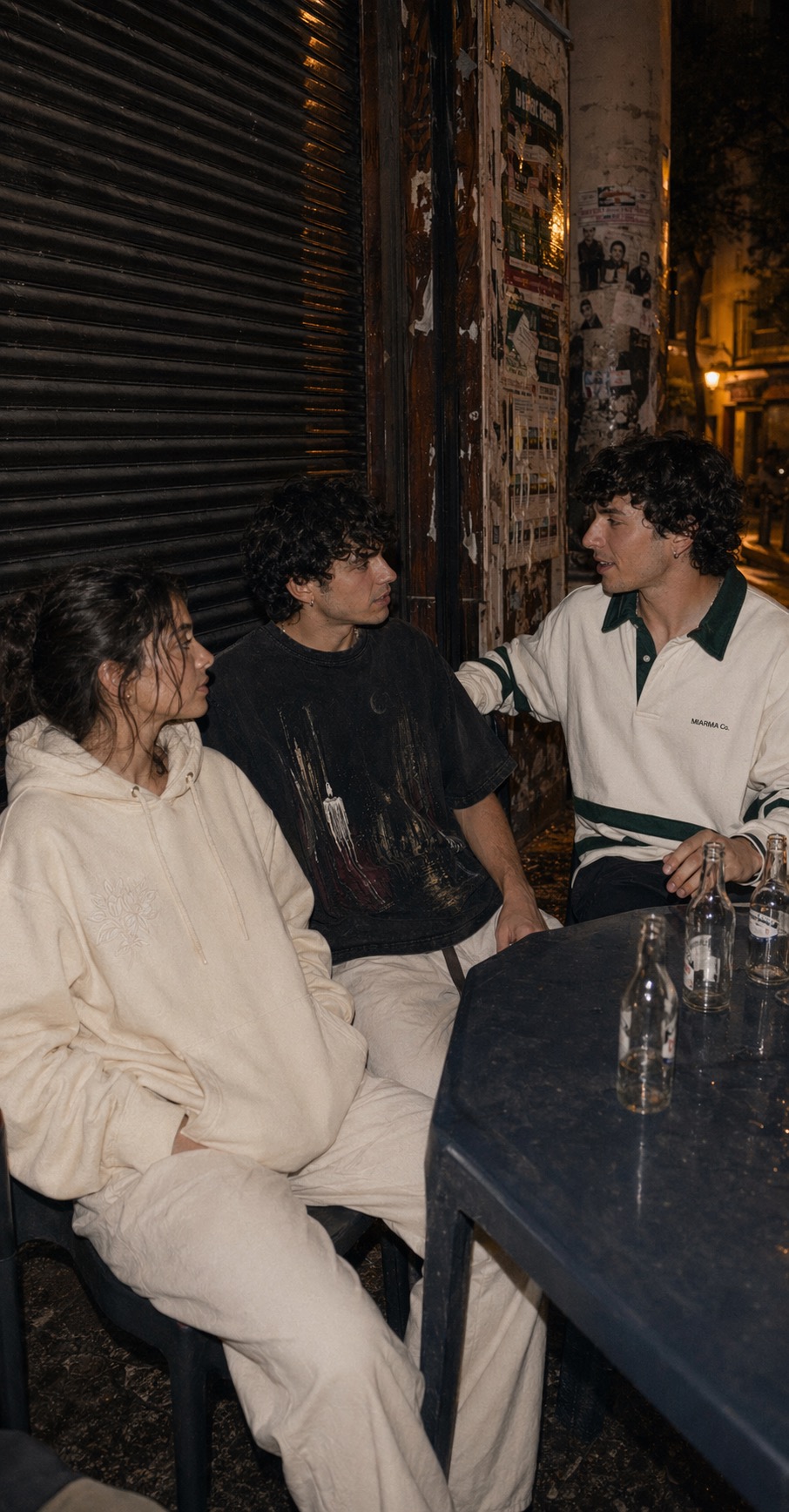Friends in the Azahar hoodie, Madruga tee, and Caseta rugby shirt sitting outside a late-night bar.