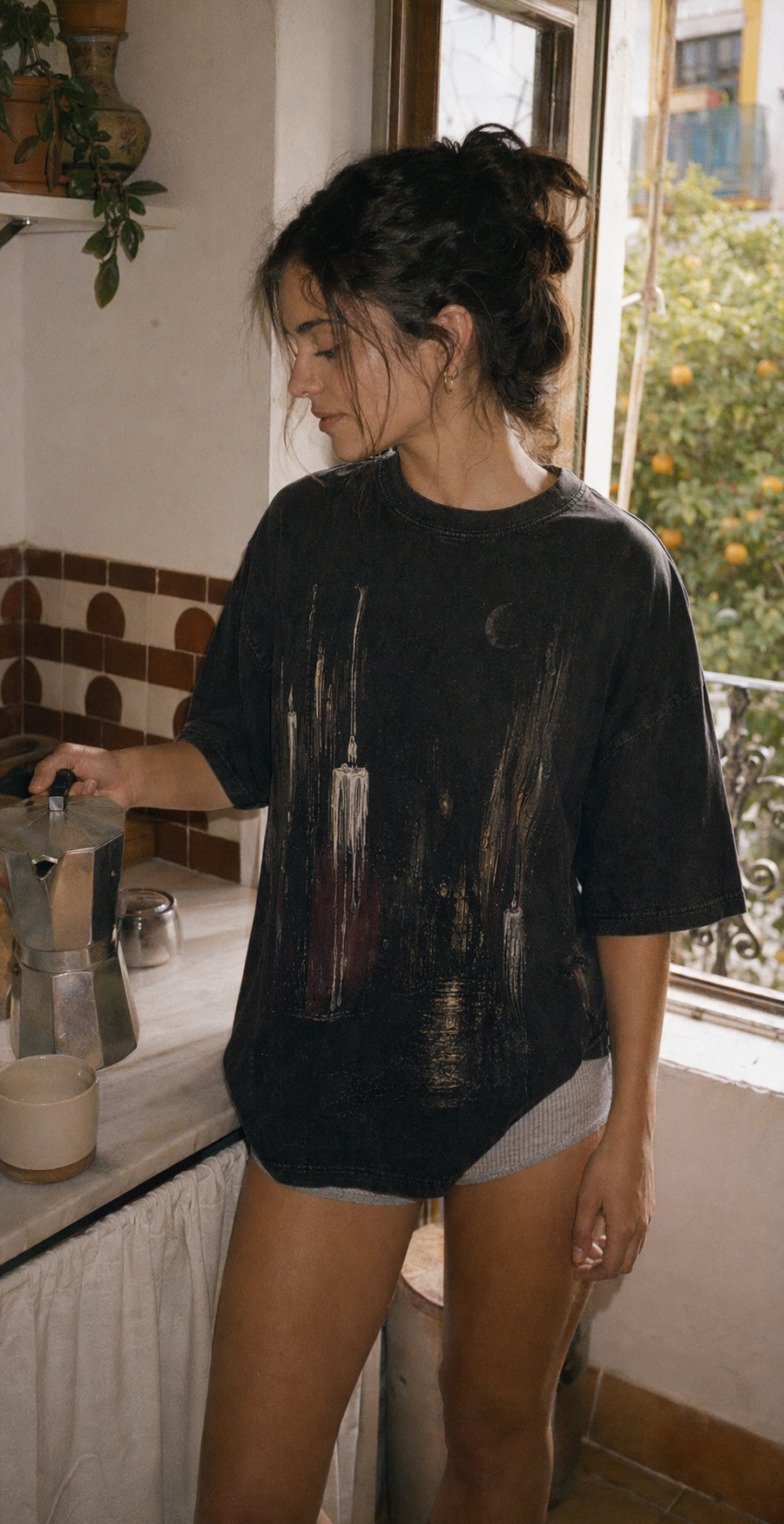 Adult woman wearing the Madruga black tee while making morning coffee in a Sevilla kitchen with orange trees outside the window.